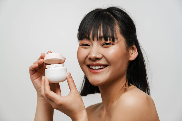 Young asian shirtless woman laughing while posing with face cream isolated over white background - Foto, Imagem