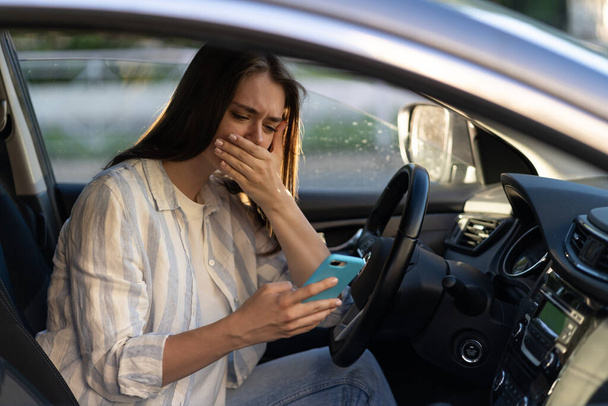 Плачущая молодая девушка читает сообщения на мобильном телефоне. Разочарованная молодая женщина на водительском сидении в машине - Фото, изображение