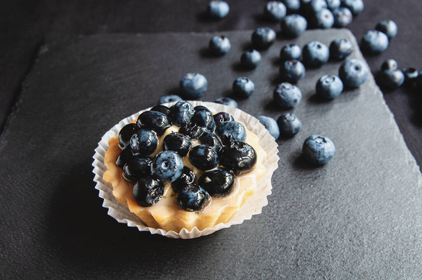 Tart with blueberries is on a black background of natural stone. Still life with sweet cakes with wild berries on a dark background - Foto, imagen