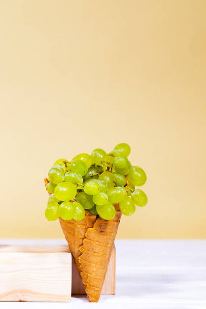 Green grape in sweet wafer cone on wooden stands over light orange background. Healthy nutrition, seasonal harvest of fruits. Copy space, Equilibrium floating food balance - Photo, Image