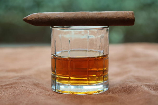 Cuban Cigar. Hand Rolled Cuban Cigar on a Glass of Whiskey on top of a Leather Table. Room for text. Single malt scotch whiskey in crystal glasses with a Cuban Cigar on Leather.  - Photo, Image