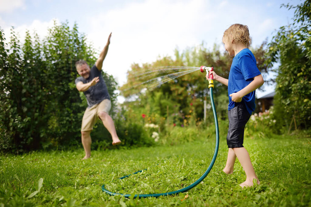 Funny little boy with his father playing with garden hose in sunny backyard. Preschooler child having fun with spray of water. Summer outdoors activity for family with kids. - 写真・画像