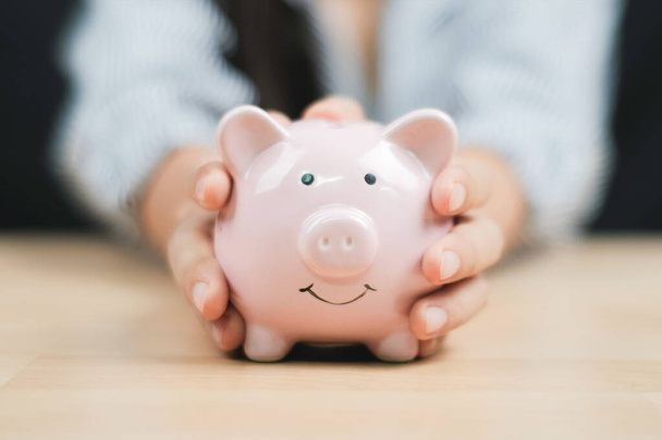Saving money concept, Women hand hold a piggy bank on wooden table. Close up.	 - Zdjęcie, obraz
