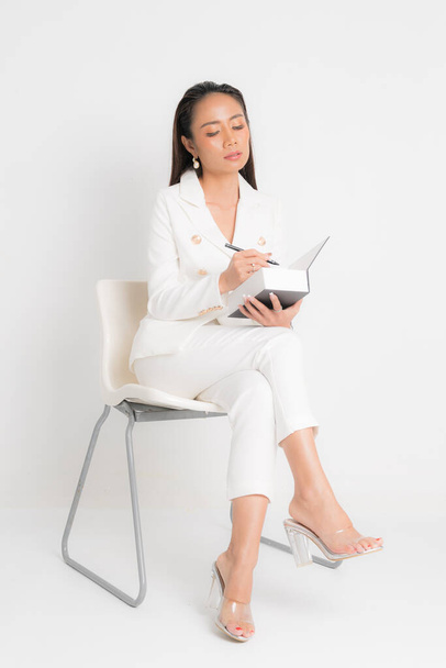 Moda styl katalog odzież dla biznesu kobieta czarny długie włosy naturalny makijaż nosić biały garnitur siedzi na białym krześle, trzymając książkę i długopisy w studio strzelać na białym tle. - Zdjęcie, obraz