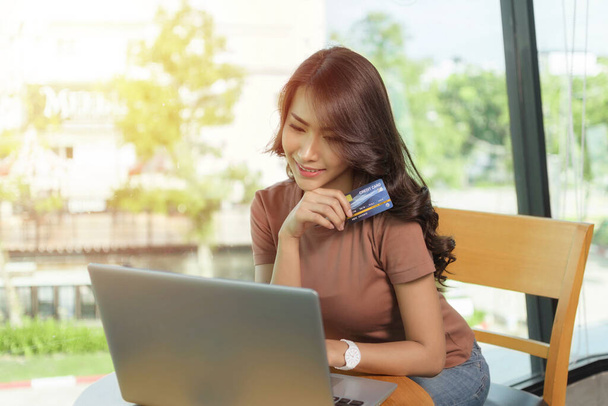 Gyönyörű nő visel barna ing ül, és mosolygós arc boldog ül egy nagy ablak, Ő vásárol online laptop és a kezében hitelkártya. - Fotó, kép