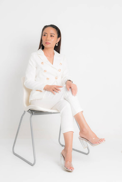 Fashion style catalogus kleding voor zakenvrouw zwart lang haar natuurlijke make-up dragen witte pak kostuum perfecte lichaam vorm pak zitten op witte stoel in studio shoot op witte achtergrond. - Foto, afbeelding