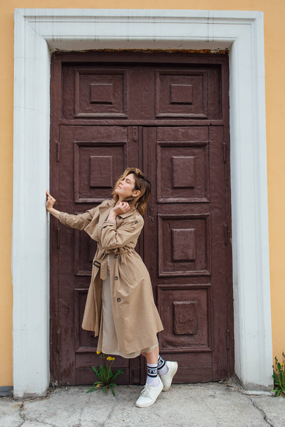 Young smiling millennial woman with wild hair dressed in an autumn coat posing near the door of an old building. - Φωτογραφία, εικόνα