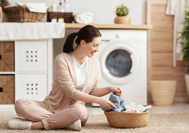 Beautiful young woman is smiling while doing laundry at home. - Zdjęcie, obraz