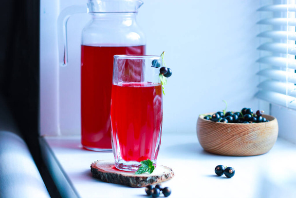 черный смородиновый сок со стаканом на окне в тени жалюзи, ягоды, летние безалкогольные напитки, здоровая пища. - Фото, изображение