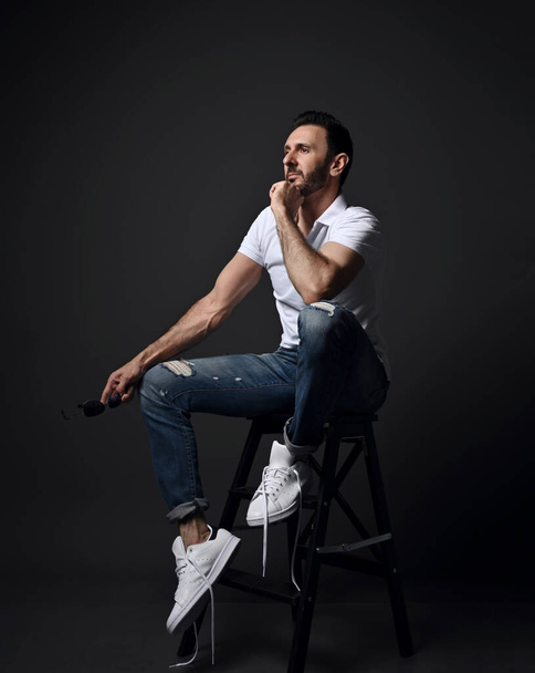 Adult unshaved man in white polo shirt, modern torn jeans and sneakers sits on high chair, stepladder looking aside - Foto, immagini