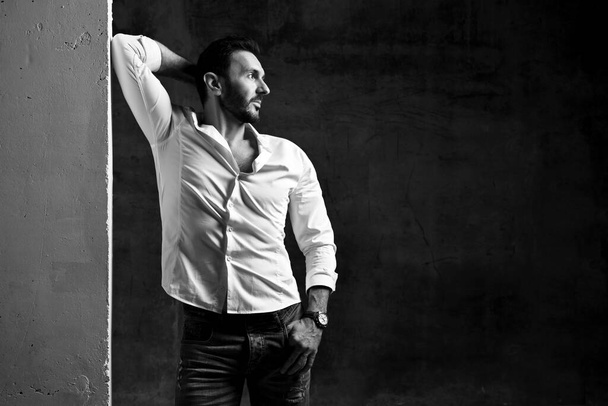 Black and white. Bearded man in white shirt and jeans leaning on concrete wall holding hand behind head looking aside - Photo, Image