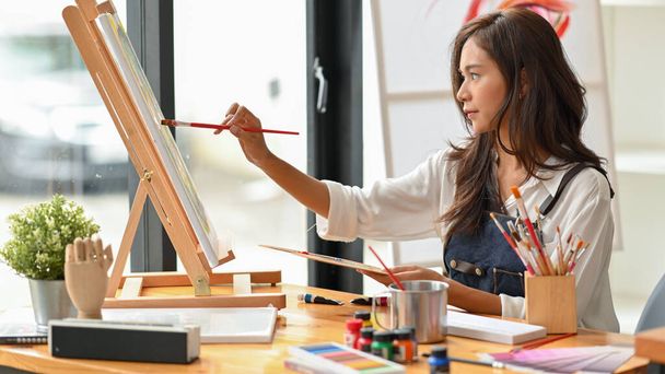 Talented female artist painting with water colour on canvas in art studio, easel, painting tools - Photo, Image