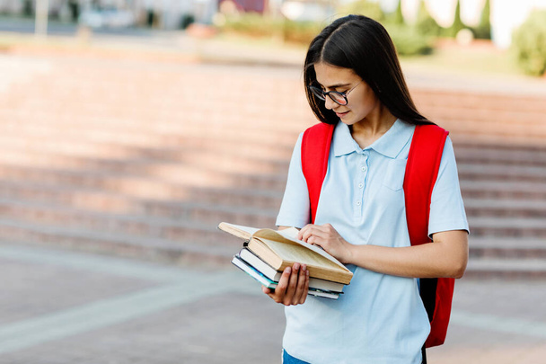 Viehättävä tyttö, jolla on reppu ja lasit, lukee kirjaa. Muotokuva opiskelijasta, jolla on kaupungin tausta. Koulutuksen käsite - Valokuva, kuva