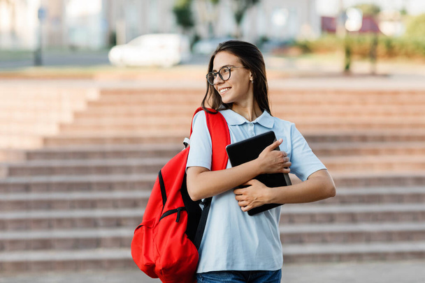 A smiling pretty student in glasses and with a red backpack, with a tablet in her hands, stands next to the stairs and smiles - Foto, afbeelding