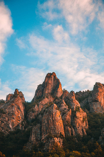 A vertical shot of rock formations on hills covered in greenery under the sunlight and a blue cloudy sky - Foto, imagen