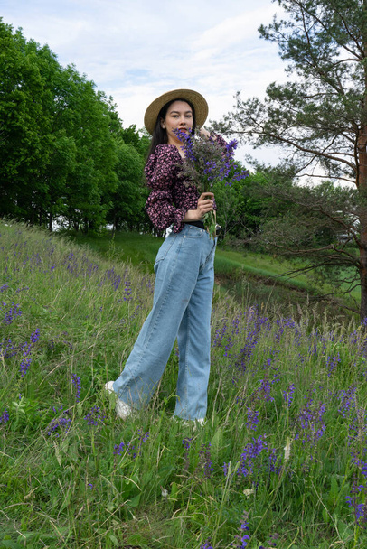 Bir buket yaban çiçeğiyle genç ve güzel bir kız dik, çiçekli bir yamaçta duruyor.. - Fotoğraf, Görsel