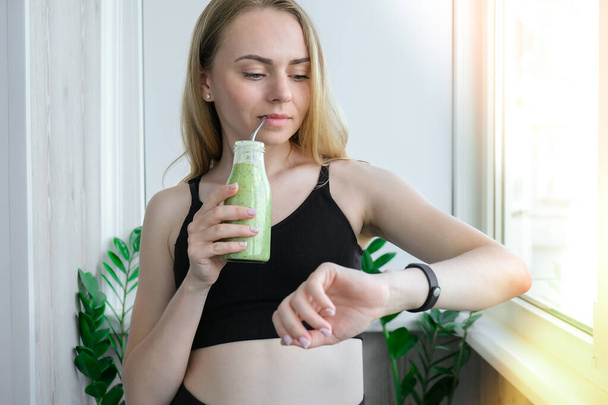 Junge Blondine trinkt veganen Smoothie-Drink Detox. Frau, die Yoga-Übungen macht, checkt Smart-Sportuhr zu Hause. Saubere Ernährung, Gewichtsverlust, gesundes Ernährungskonzept. Bleiben Sie gesund. Wellness - Foto, Bild