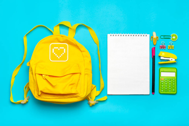 Zurück zur Schule, zum Bildungskonzept. Gelber Rucksack mit Schulmaterial - Notizbuch, Stifte, Lineal, Taschenrechner, Schere isoliert auf blauem Hintergrund - Foto, Bild