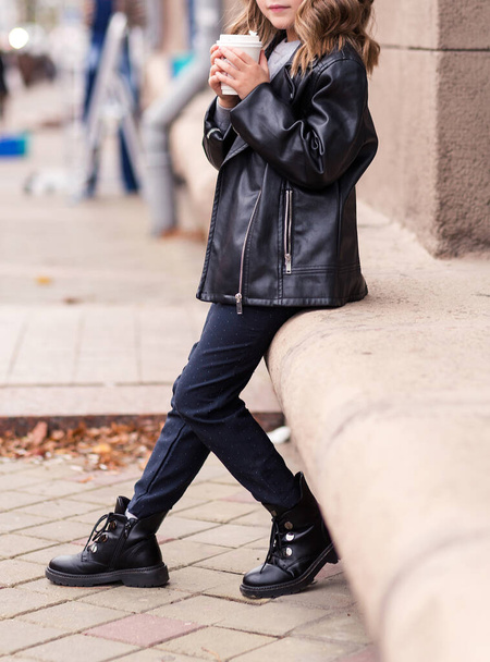 Κομψό κορίτσι με μαύρο δερμάτινο μπουφάν και μαύρο παντελόνι που ποζάρει σε δρόμο της πόλης με ένα ποτήρι ζεστό ποτό στα χέρια της. Φωτογραφία - Φωτογραφία, εικόνα