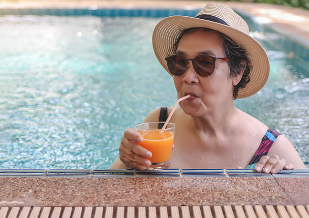 Ritratto di donna anziana asiatica felice e sana con cappello di paglia e occhiali da sole, che beve succo d'arancia in piscina, sorride e guarda la macchina fotografica. Stile di vita attivo e concetto di viaggio. - Foto, immagini
