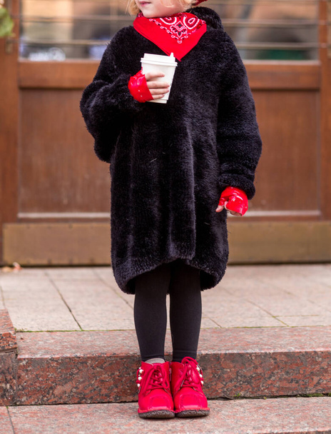 Sonbahar havasında elinde bir bardak sıcak çikolatayla şehir caddesinde poz veren siyah ceketli, kırmızı kaşkollü ve botlu bir kız. Fotoğraf. - Fotoğraf, Görsel