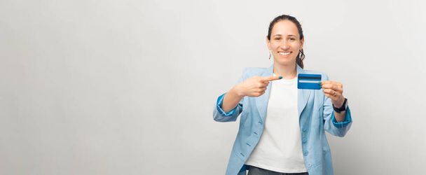 Cheerful lady smiling at the camera is pointing at a credit card over white background. - Photo, Image