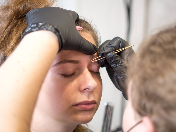 Постоянный макияж. Красивая молодая женщина проходит процедуру коррекции бровей. Выщипывание бровей девушки пинцетом - Фото, изображение