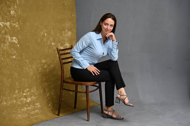 High resolution full length portrait of smiling young woman in blue shirt and trousers posing while sitting on chair against beige and gray background - Foto, Bild