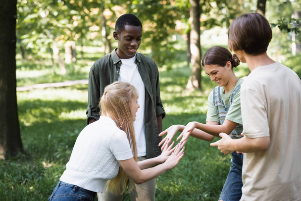 Uśmiechnięte nastolatki bawiące się w pobliżu wielonarodowych przyjaciół w parku  - Zdjęcie, obraz