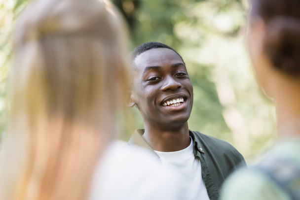 Χαμογελώντας Αφροαμερικανός έφηβος κοιτάζοντας την κάμερα κοντά σε θολή φίλους  - Φωτογραφία, εικόνα