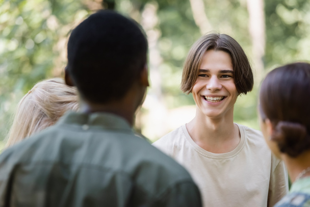Smiling teenager looking at blurred interracial friends outdoors  - Fotó, kép