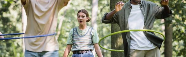 Excited teen girl looking at camera near interracial friends with hula hoops in park, banner  - Photo, Image