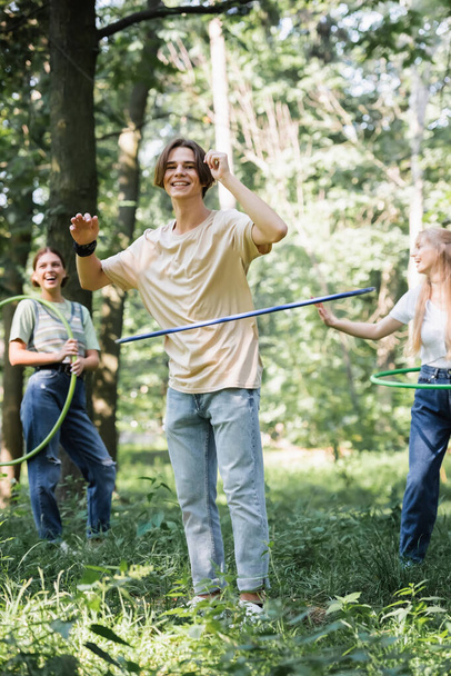 Smiling teenager twisting hula hoop near cheerful friends on blurred background in park  - Foto, Imagen
