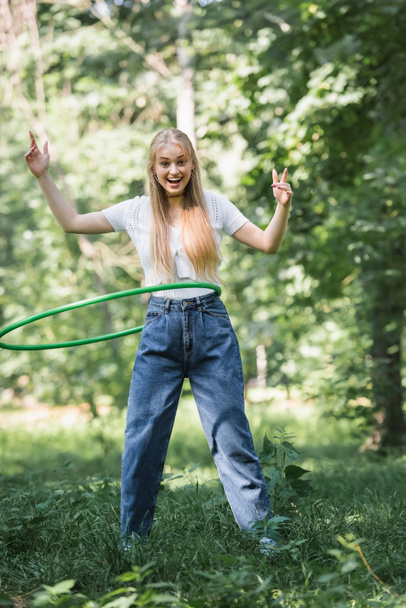 Sonriente adolescente torciendo hula hoop y mirando a la cámara en el parque  - Foto, imagen
