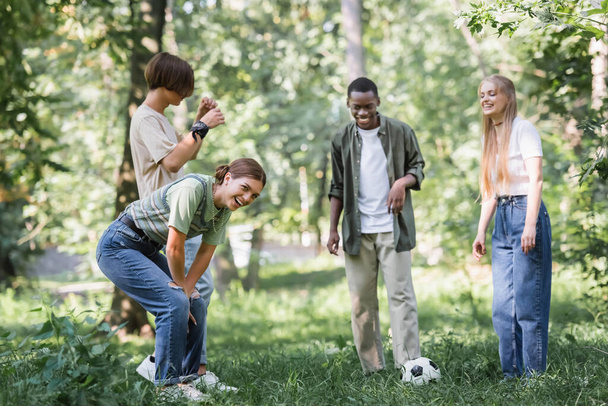 Adolescente sorrindo olhando para a câmera perto de futebol e amigos inter-raciais no parque  - Foto, Imagem