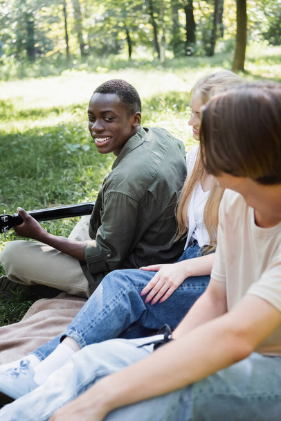 glimlachend afrikaanse tiener spelen akoestische gitaar in de buurt wazig vrienden in park  - Foto, afbeelding