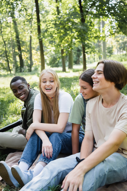 Cheerful teen friends sitting near african american boy with acoustic guitar in park  - Valokuva, kuva