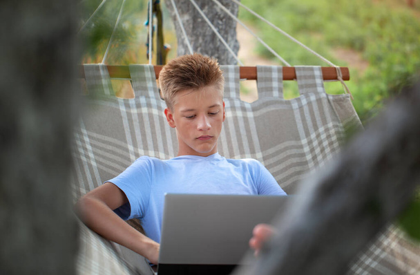Adorable young teenage man with a laptop lying in hammock in summer garden. Concept distant work, education, freelancer, rest and relaxation - Foto, imagen