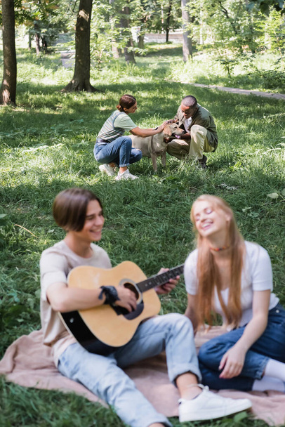 Interracial teenagers petting retriever near blurred friends playing acoustic guitar on lawn  - 写真・画像