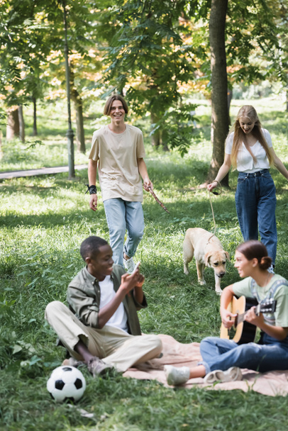 Uśmiechnięte nastolatki rozmawiające w pobliżu retrievera i rozmyty multietniczny przyjaciel z gitarą akustyczną na trawie  - Zdjęcie, obraz