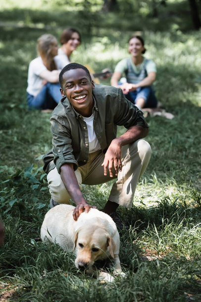 Sonriente afroamericano adolescente mirando a la cámara cerca de retriever en el césped  - Foto, imagen