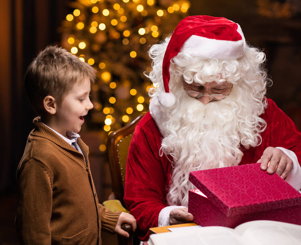 Санта Клаус і маленький хлопчик. Веселий Санта працює, сидячи за столом. Вогонь і різдвяне дерево на задньому плані. Різдвяна концепція. - Фото, зображення
