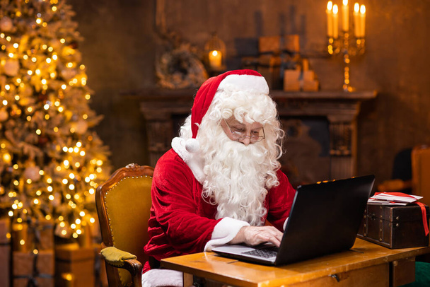 Workplace of Santa Claus. Cheerful Santa is working on the laptop while sitting at the table. Fireplace and Christmas Tree in the background. Christmas concept. - Photo, Image