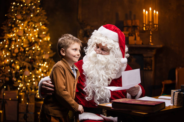 Santa Claus and little boy. Cheerful Santa is working while sitting at the table. Fireplace and Christmas Tree in the background. Christmas concept. - Foto, imagen