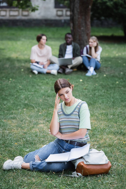 Плотный студент с блокнотом и рюкзаком сидит рядом с размытыми друзьями на улице - Фото, изображение