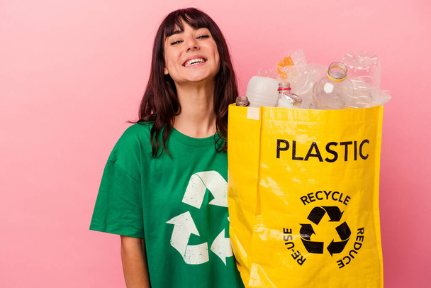 Junge kaukasische Frau hält eine recycelte Plastiktüte isoliert auf rosa Hintergrund lachen und Spaß haben. - Foto, Bild