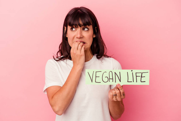 Giovane donna caucasica in possesso di un cartello di vita vegana isolato su sfondo rosa mordere le unghie, nervoso e molto ansioso. - Foto, immagini