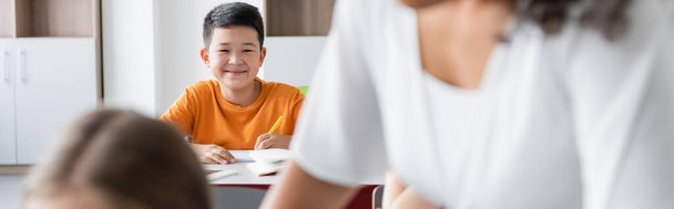cheerful asian schoolboy looking at camera near teacher on blurred foreground, banner - Foto, afbeelding