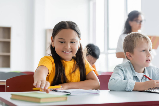 fröhliche asiatische Schülerin greift nach Buch, während sie am Schreibtisch neben Klassenkameradin sitzt - Foto, Bild