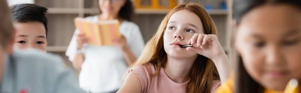fille coûteuse tenant stylo dans la bouche tout en regardant loin près floue élèves multiethniques, bannière - Photo, image